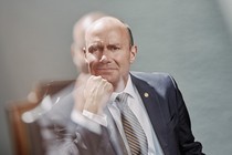 A color portrait of Mike Lee in suit and tie, sitting in chair with hand to chin, with double exposure of image