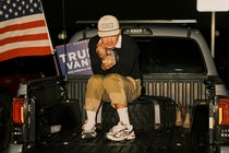 Young man sitting in the back of a pickup truck, checking his phone, with a Trump campaign sign and the American flag behind him
