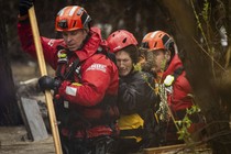 Two rescue workers help a person escape a flood, walking through waist-deep water.