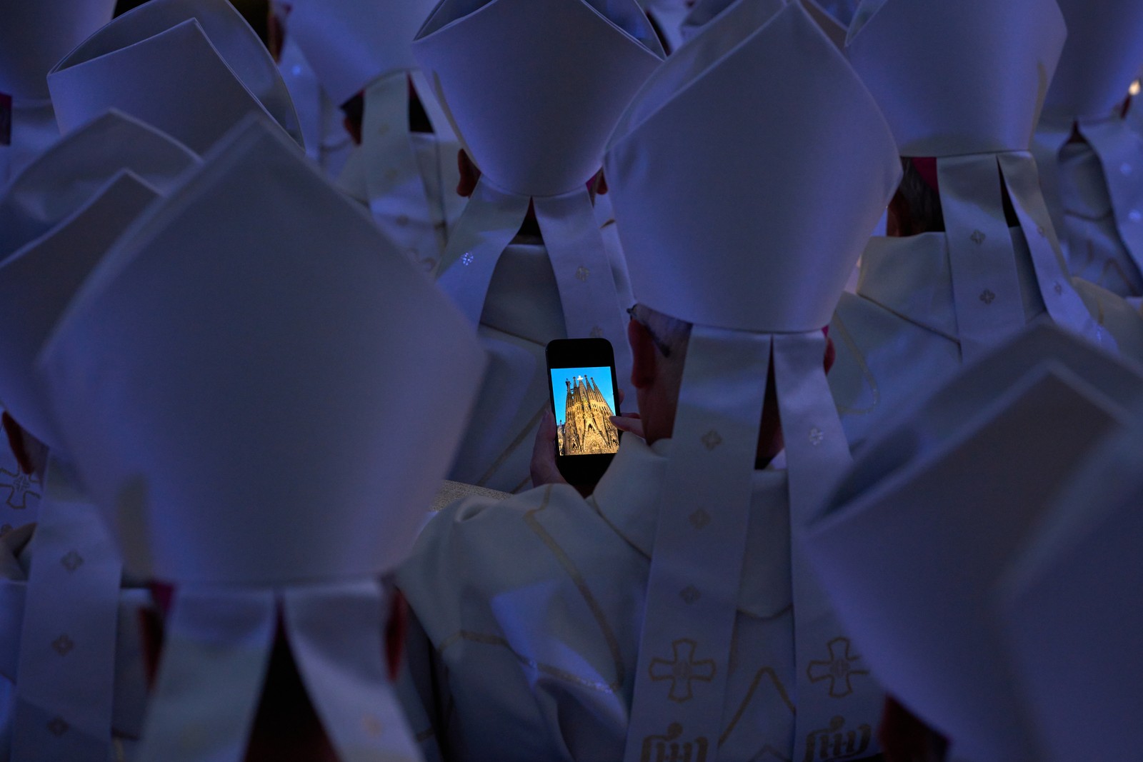Priests in ceremonial gear gather during a ceremony, with one holding a cell phone that displays a photograph of a cathedral.