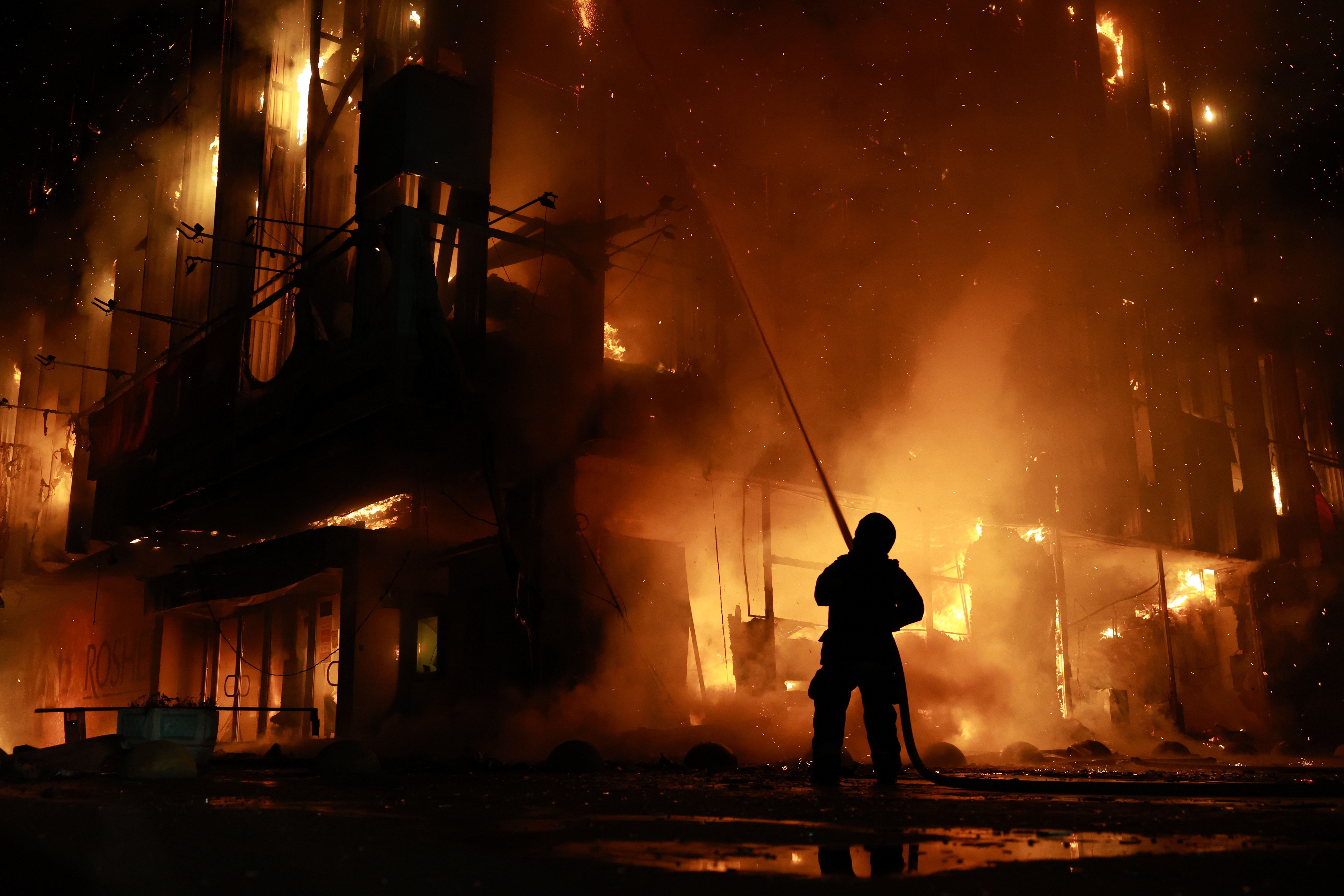 A person uses a hose to spray water onto a tall burning building at night.