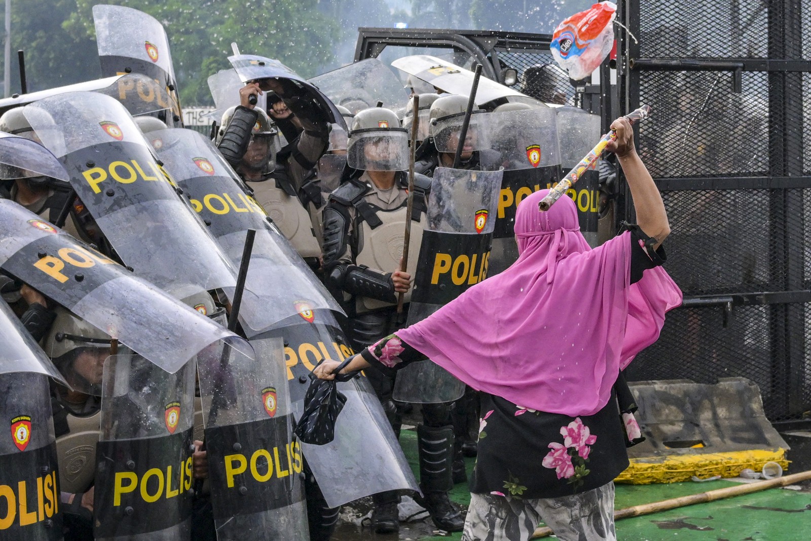 A protester swings a bamboo stick toward a group of riot police officers holding shields.