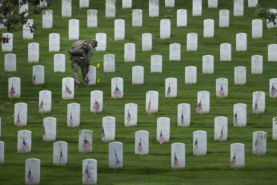 A soldier places small American flags beside dozens of headstones.
