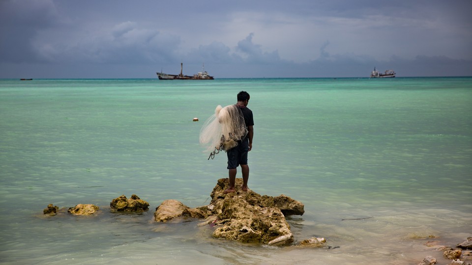 Kaitara Kautu, a net fisherman whose home flooded during last year's king tide in Betio, a town on the island of South Tarawa in Kiribati, stands on a rock in the middle of turquoise-colored water; a net rests on his left shoulder, and large ships sit in the distance.