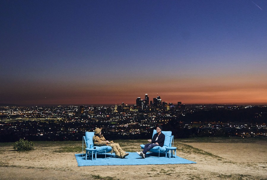 Two people sit in chairs outside at sunset, in front of the Los Angeles skyline.