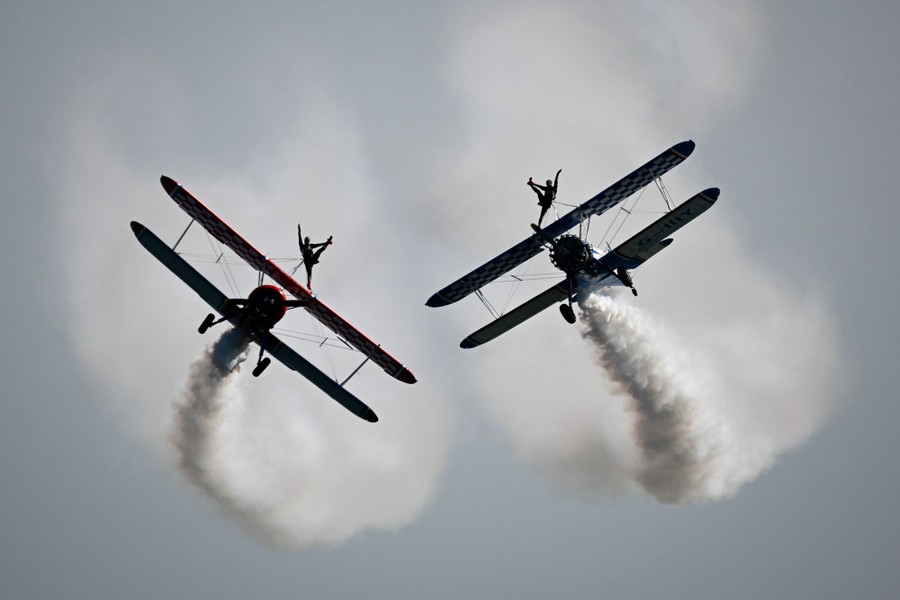 Two acrobats stand (strapped in) atop biplane wings as the aircraft fly close to each other.