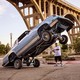 photo of hydraulic car resting on back wheels with front wheels in air above the head of man standing behind it, with tall arched bridge in background