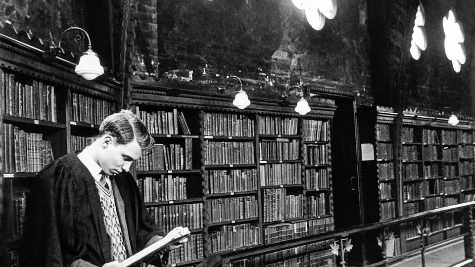 A black and white photograph of a student reading a book on the upper level of a library, leaning against a balcony