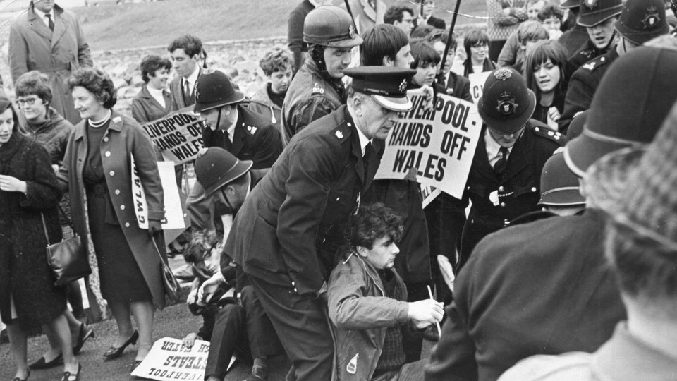 Welsh-nationalist demonstrators holding signs that read "Liverpool—hands off Wales" are dragged away by police officers.