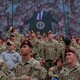 A group of soldiers stands in front of a Jumbotron showing Donald Trump at a podium above them