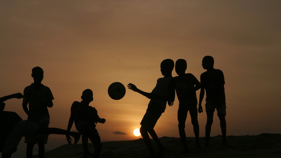 The silhouettes of a group of children kicking a soccer ball in front of the setting sun