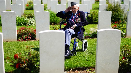 An elderly veteran wearing many medals sits in a wheelchair, saluting, among many rows of headstones in a cemetery.
