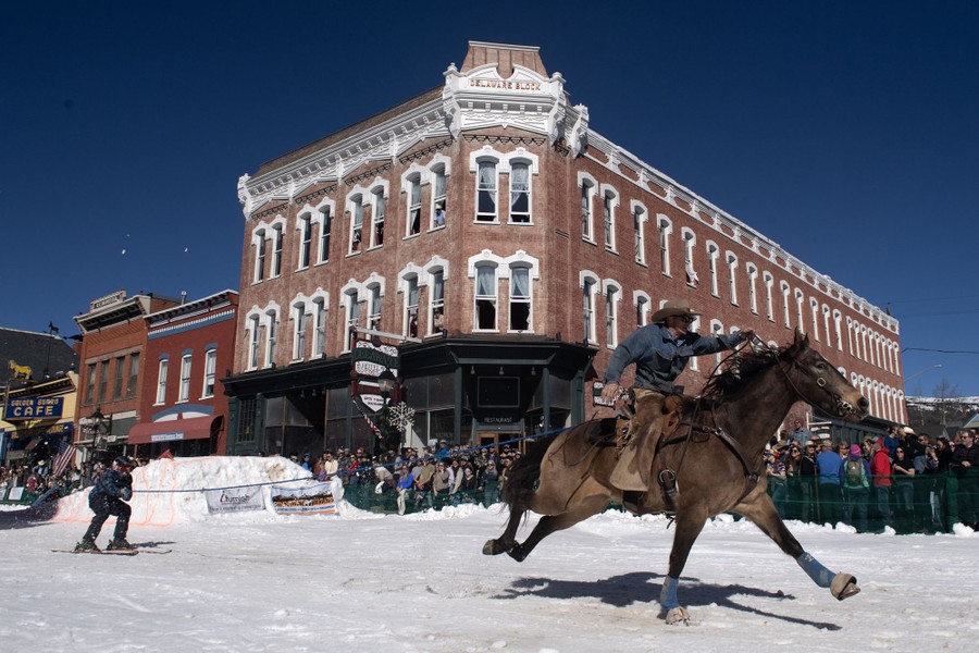 A rider on a horse pulls a skier with a tow rope down a snow-covered city street, in front of a crowd.