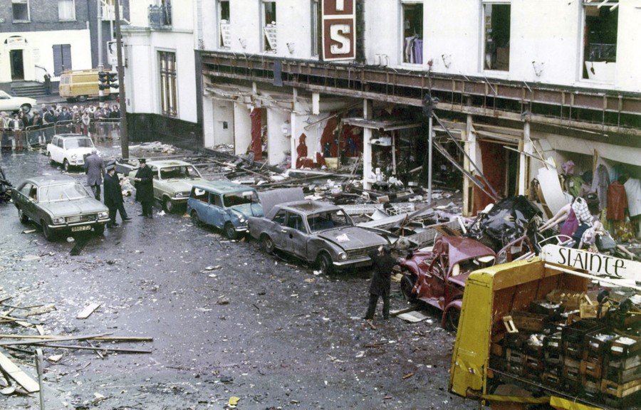 Damaged cars sit covered in debris in a street beside a bomb-damaged building.