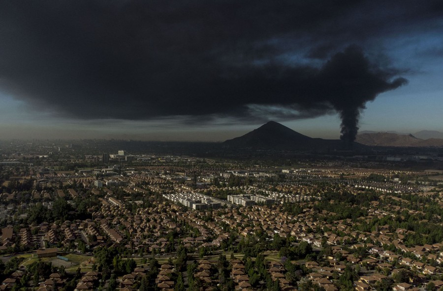 A view looks across houses and buildings; a large column of black smoke rises from a fire in the distance.