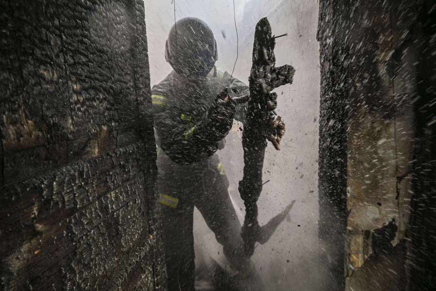 A firefighter walking through a doorway in a burning building with water and debris flying around.