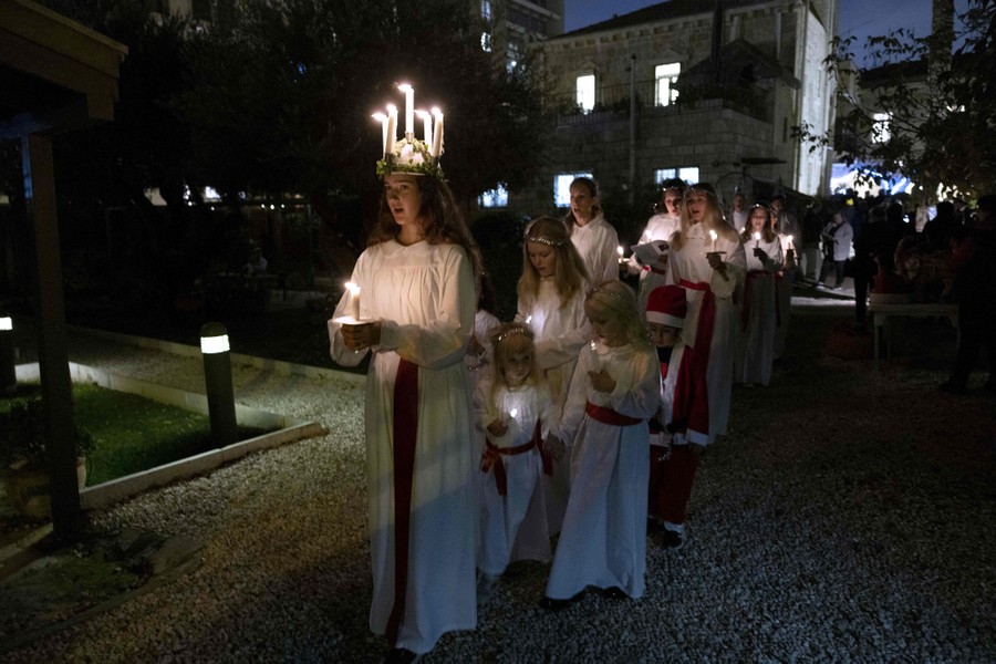 A procession of young people dressed in white walks behind a young woman who wears a crown of candles.
