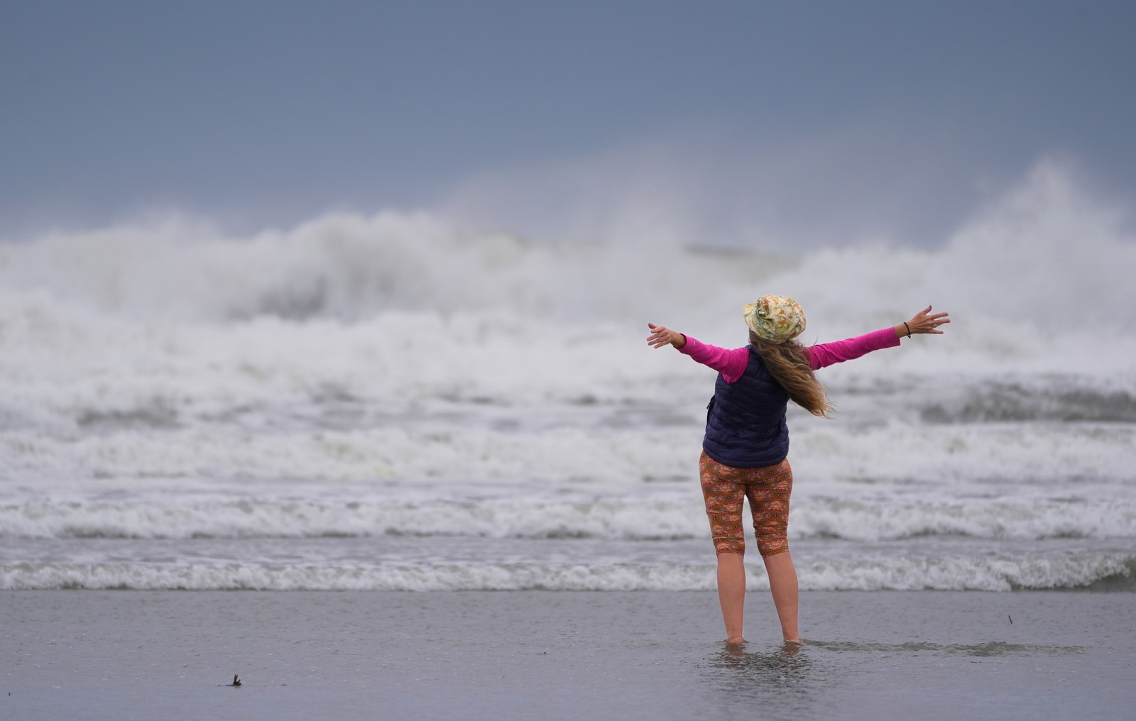 A person stands on a beach during a storm, facing the rough water, with arms wide open.