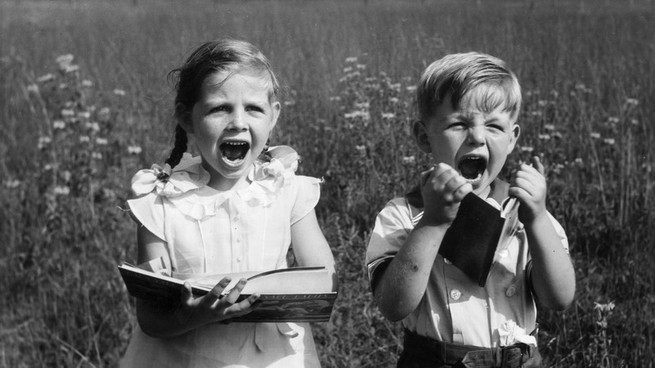 Two children in a field display extreme emotion while holding books.