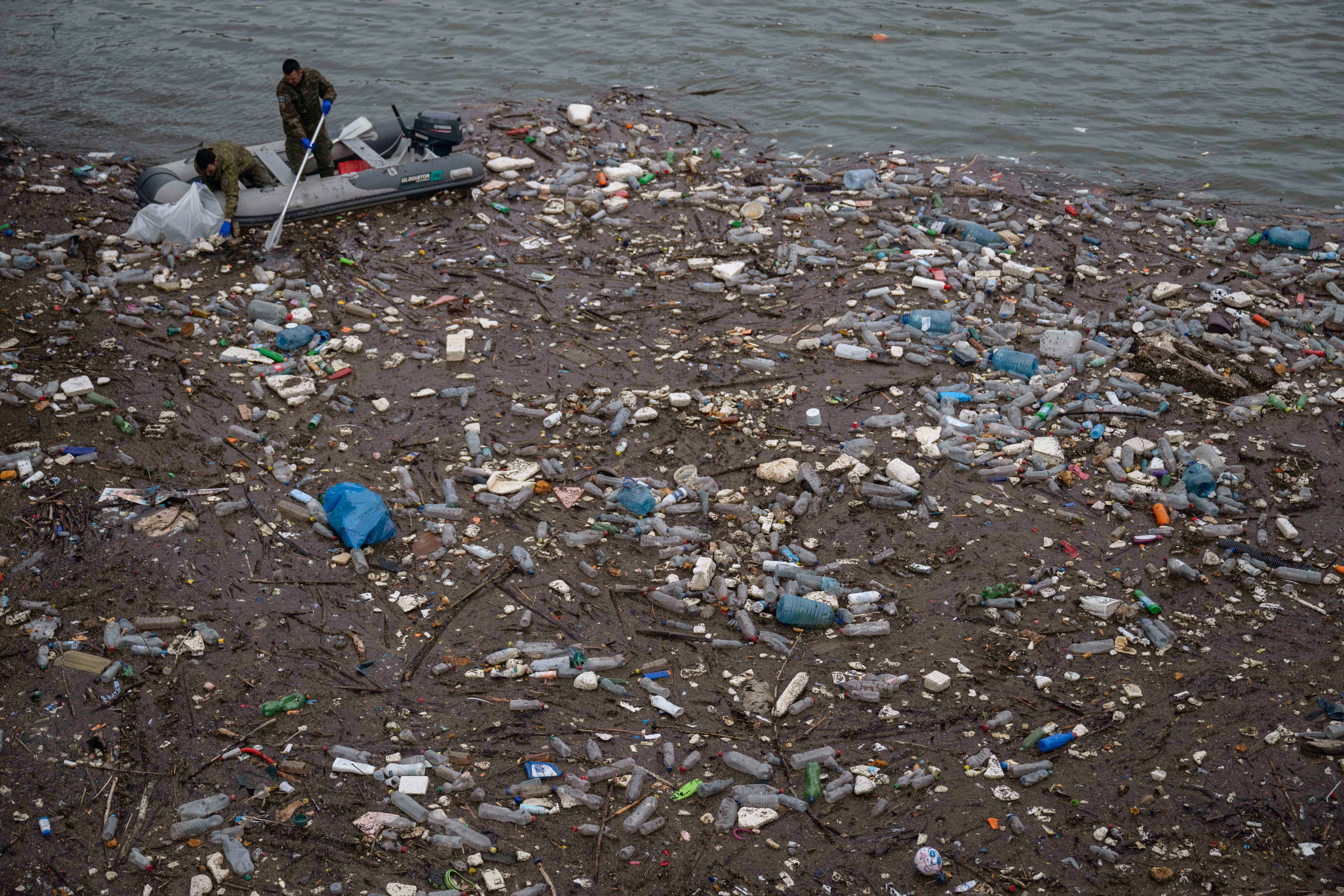 Two people in an inflatable boat work to clear debris from a large patch of floating garbage in a lake.