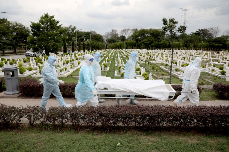 Five people wearing personal protective equipment carry a stretcher with a covered body on it through a cemetery.