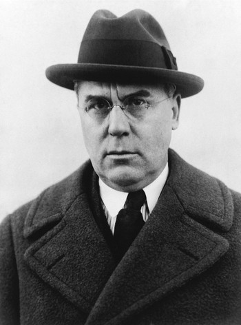 black-and-white photo of man wearing fedora, glasses, and heavy overcoat over shirt and tie