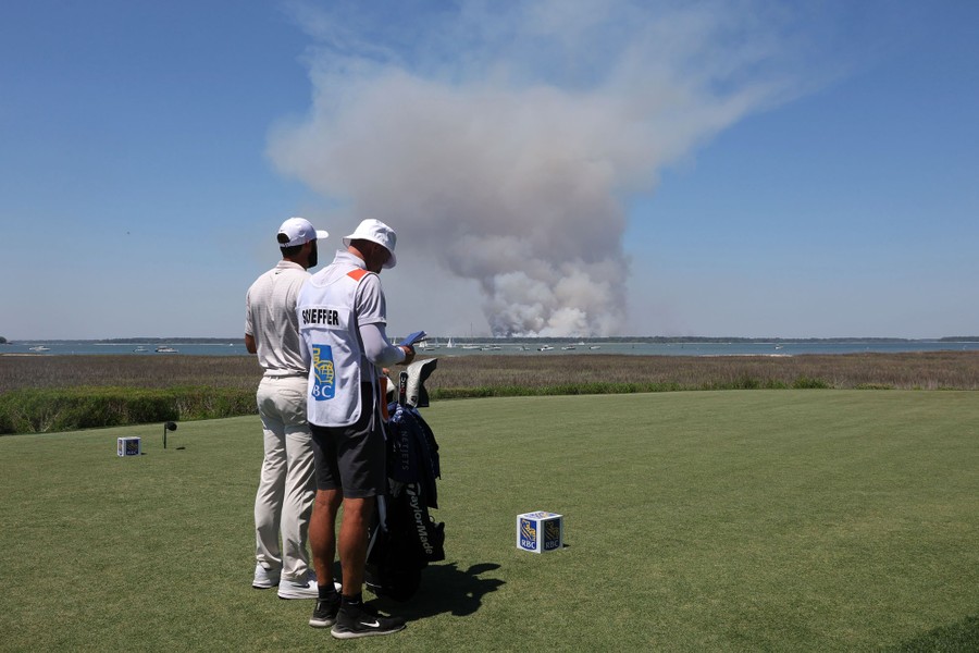 A golfer and caddy stand at a golf tee, with smoke from a brush fire rising in the distance.