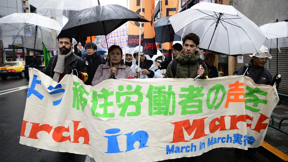 Migrant workers march together on the streets of Tokyo holding banners and umbrellas.