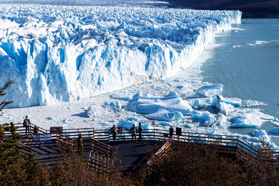 Half a dozen people stand in a viewing area, looking out toward a large glacier.