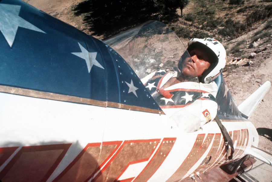 A stunt performer sits in the cockpit of a small rocket ship, tilted up at a steep angle.