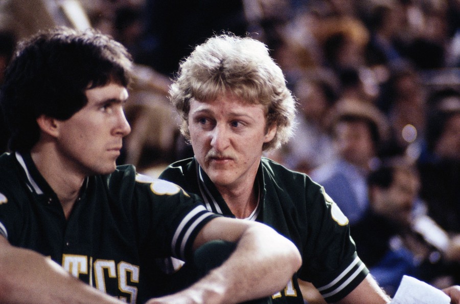 A portrait of Larry Bird sitting on the sidelines of a basketball game.