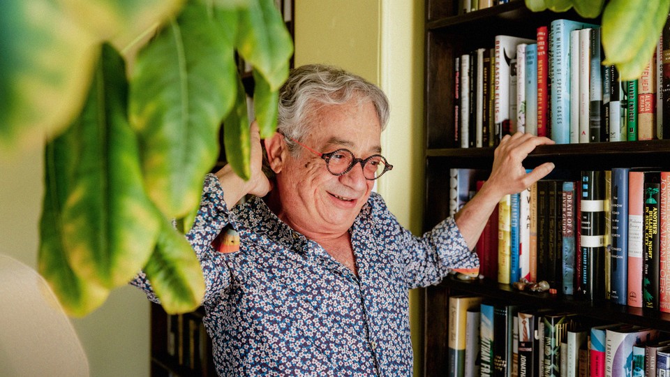 Portrait of Rabih Alameddine standing by a bookshelf with the hanging leaves of a tree in the foreground