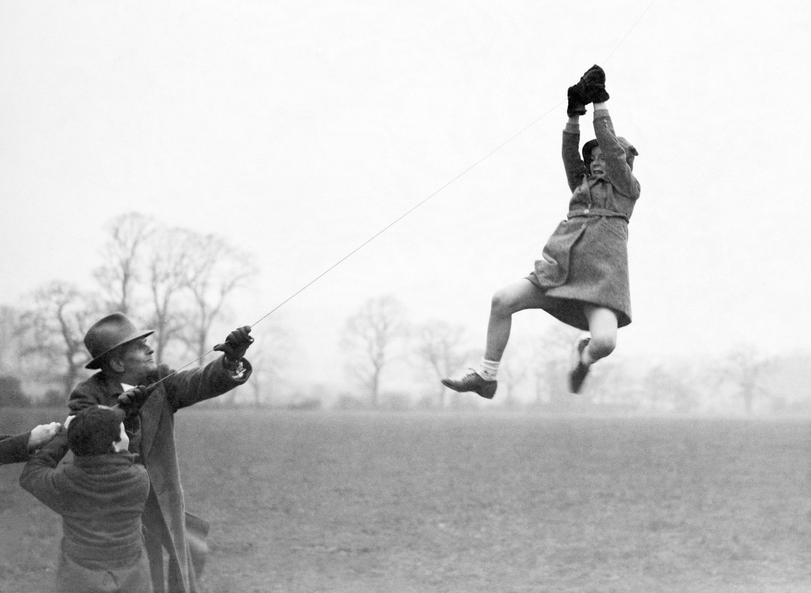 A man and boy flying a large kite work to help a girl that has been lifted off her feet and is hanging onto the kite string.