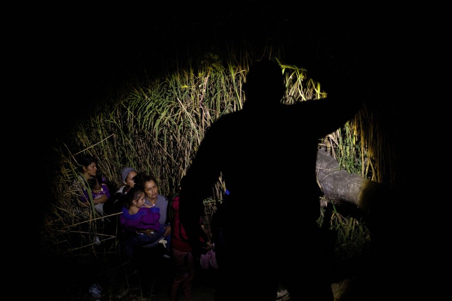 Several people are seen walking through tall grass at night, illuminated by a flashlight held by a Border Patrol agent seen in silhouette.