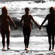 Three nude sisters hold hands on the beach.