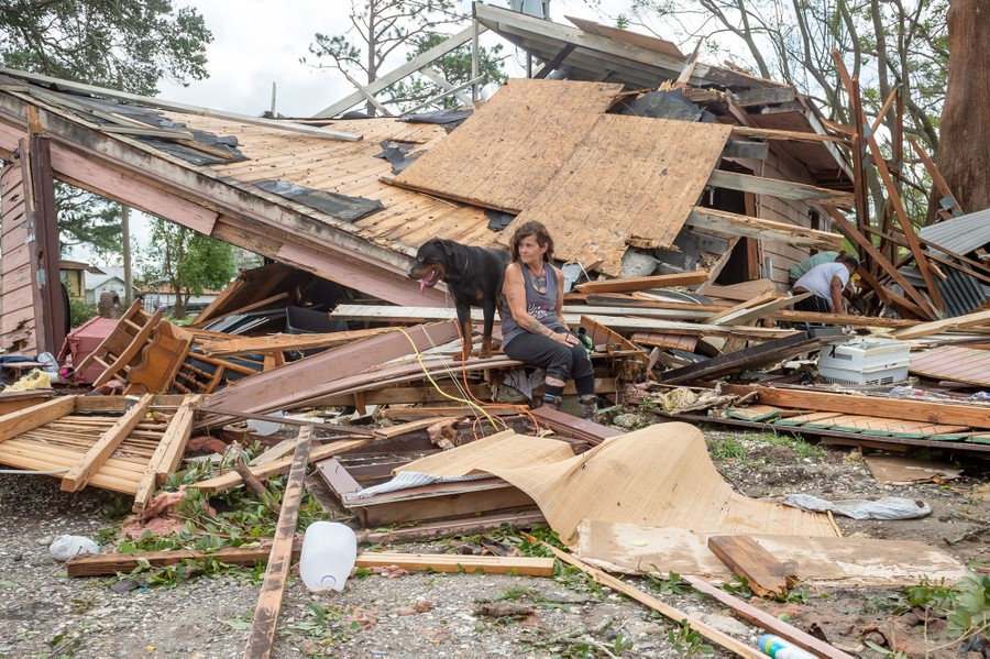 A woman and her dog sit on a piece of debris in front of her destroyed home.