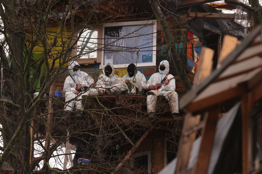 Four people wearing white protective gear sit on a ledge outside a treehouse.
