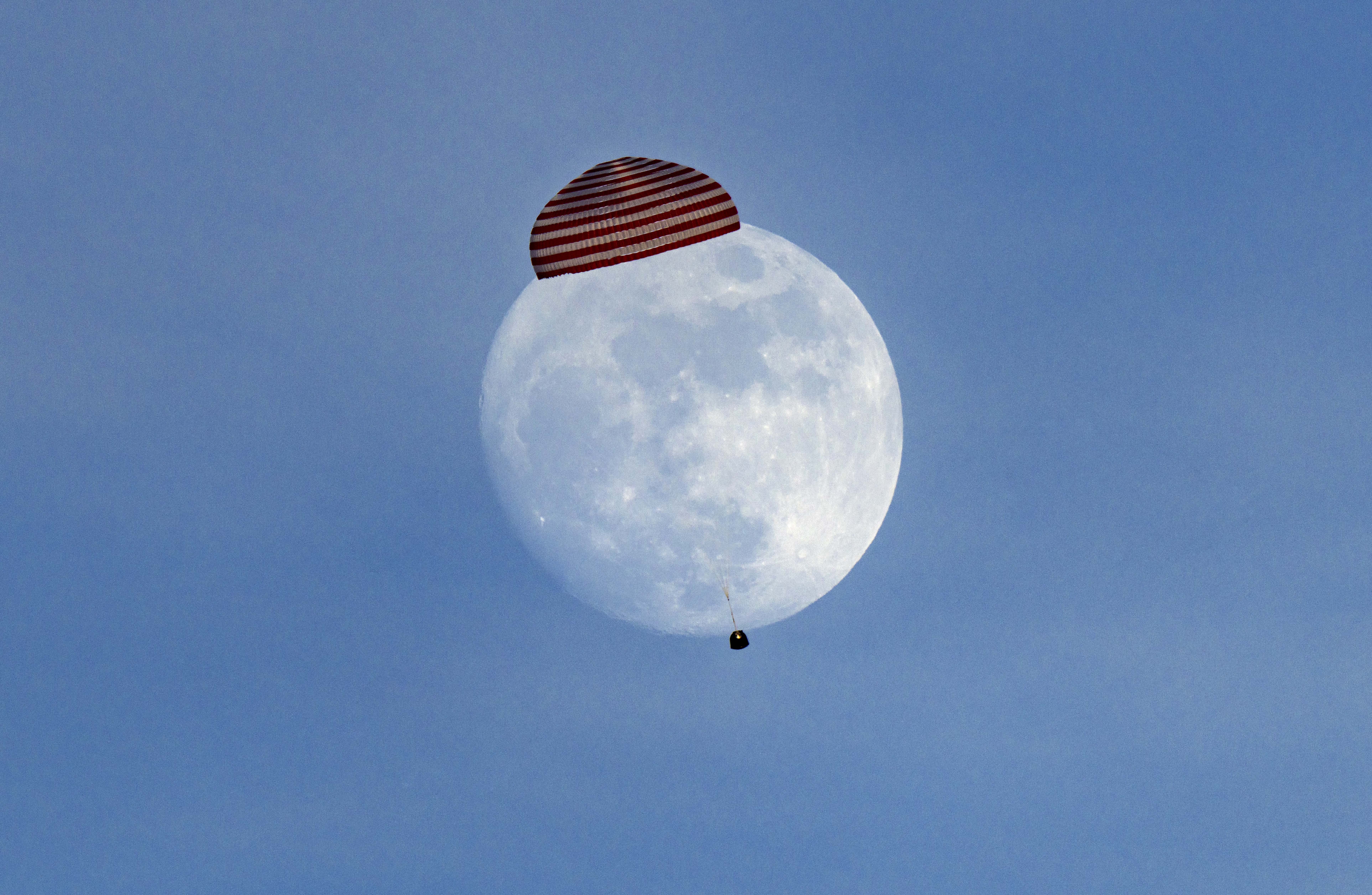 A space capsule descends under a parachute, framing a distant full moon.