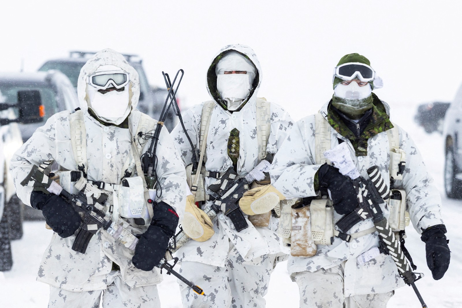 Three soldiers walk down a road in a snow-covered area. All wear white camouflage uniforms and carry weapons.
