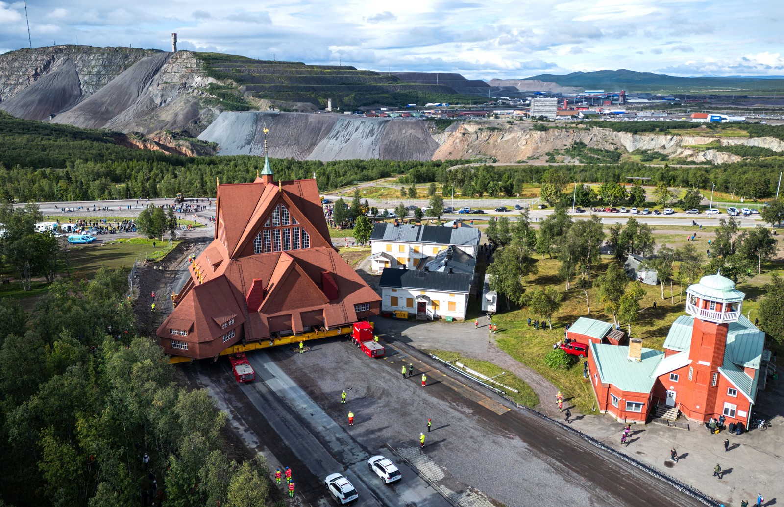 A tall wooden church, supported by steel beams mounted atop wheeled tractors moves slowly down a road, seen from above.