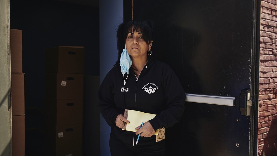 A woman, dressed in a black track suit and with a blue surgical mask hanging off one ear, stands in a doorway.
