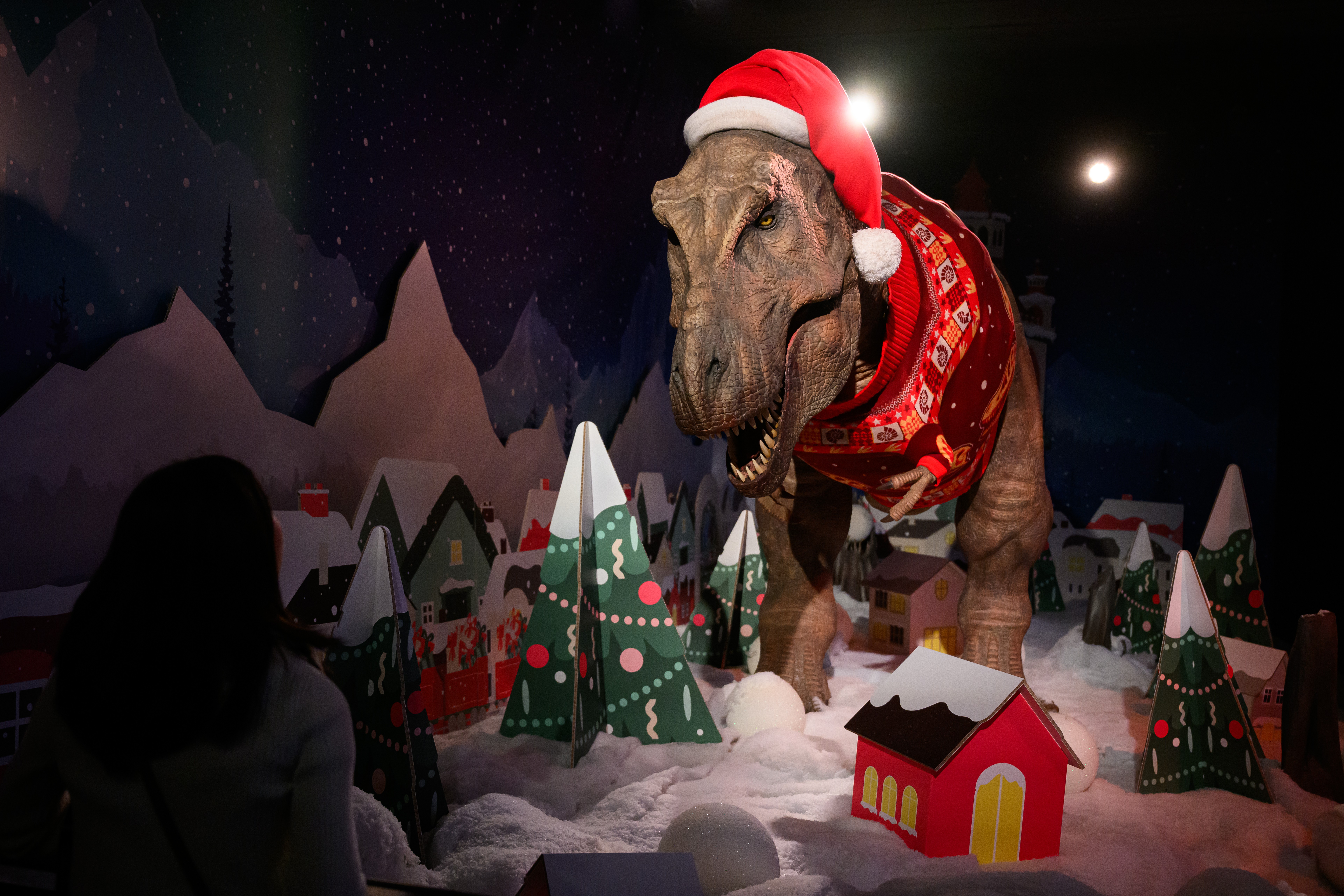 A large Tyrannosaurus Rex statue, wearing a Christmas sweater, in a holiday display.