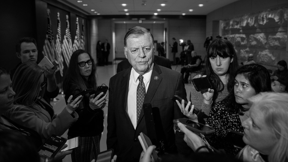 Black-and-white photograph of Tom Cole, dressed in a suit, surrounded by reporters holding their phones up toward him.