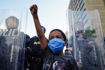 A young black boy wearing a mask raises his fist in front of a line of police officers using riot shields