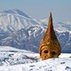 An old gold-colored statue of a head with a tall hat sits on snow-covered ground with mountains in the background.