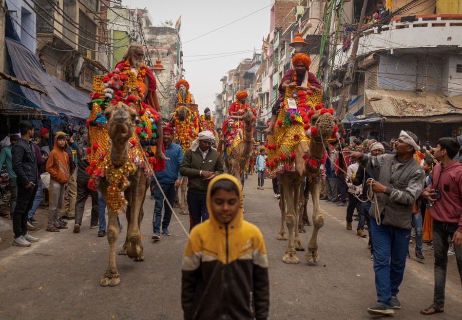Several holy men draped in colorful clothing and flowers ride atop camels, being led down a city street.