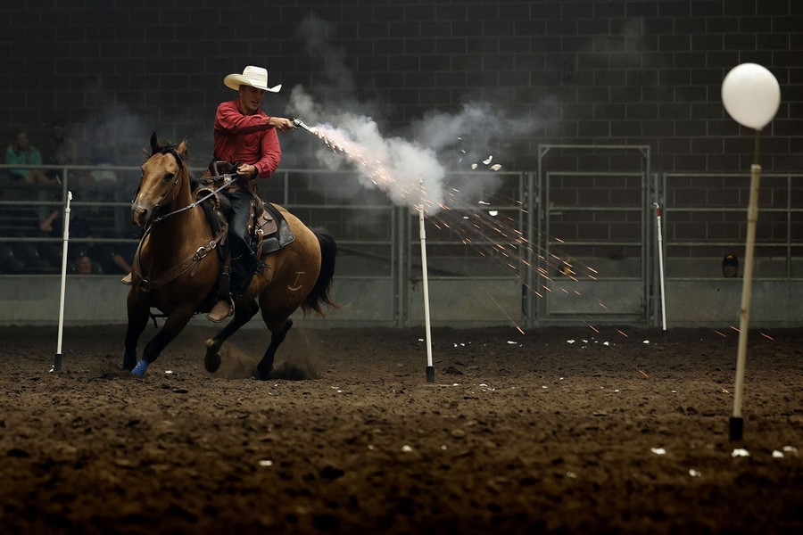 A rider on horseback, inside a stadium, fires a pistol at balloon targets on sticks.