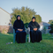 two women in hijabs and long-sleeved black dresses sit, hands in laps, in lawn chairs on green grass in yard next to house