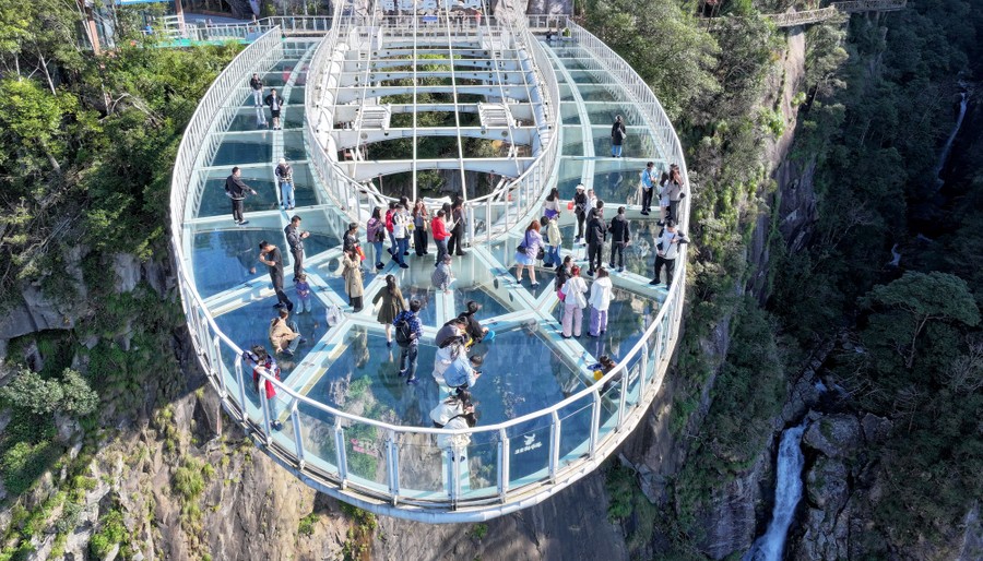 A group of visitors walk on a cantilevered, glass-floored observation deck above a cliff.