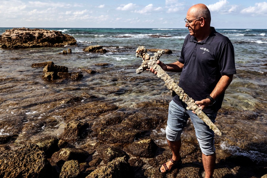 A person stands on a rocky seashore, holding an ancient sword encrusted with shells that was recovered from the ocean.